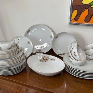 Könnte beinhalten: Ein komplettes weißes Porzellan-Geschirrset mit Goldrand und zartem Blumendesign. Das Set beinhaltet Teller, Untertassen, Tassen, eine Sauciere und eine Servierschüssel. Das Geschirr ist auf einer Holzoberfläche angeordnet.