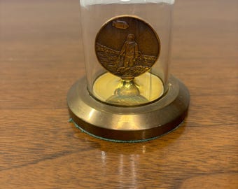 May include: A bronze medallion depicting an astronaut, housed within a glass dome. The dome rests on a brass base with a green felt underside. The astronaut is shown on a textured surface, possibly the moon.