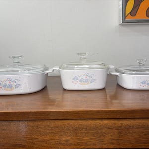 May include: Three white casserole dishes with glass lids. Each dish features a floral design with blue and yellow accents. The dishes are arranged on a wooden surface, showcasing their varying sizes.