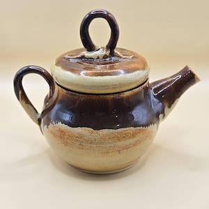 May include: A ceramic teapot with a lid and handle. The teapot is mainly brown with a lighter tan band around the middle. The handle and spout are dark brown. The lid has a circular handle.