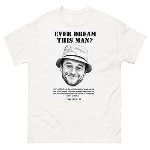 Könnte beinhalten: Ein weißes kurzärmeliges T-Shirt mit einer schwarz-weißen Grafik einer Person mit Fischerhut. Über der Grafik steht der Text "EVER DREAM THIS MAN?" und darunter ein Text über Träume mit der Telefonnummer (254) 231-8732.