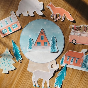 May include: A set of colorful paper cutouts featuring a snow globe with a cabin inside, a polar bear, a fox, a deer, a Christmas tree, and two houses. The snow globe is blue and white with snowflakes. The cabin is red with a white roof and a wreath on the door. The polar bear is white and the fox is orange. The deer is brown and white. The Christmas tree is green with white snowflakes. The houses are red with white trim.