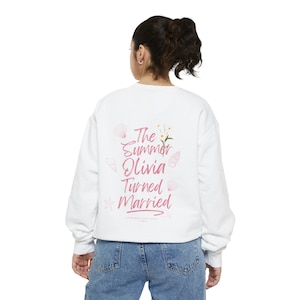 Peut inclure: Sweat-shirt blanc avec un motif rose qui dit "The Summer Olivia Turned Married" avec des coquillages et des fleurs.