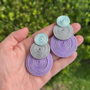 Pode incluir: Um par de brincos pendentes com tr&ecirc;s discos circulares empilhados de tamanhos variados. Cada disco &eacute; constru&iacute;do com fio enrolado firmemente em azul claro, prata e roxo, dispostos num padr&atilde;o vertical do menor para o maior.