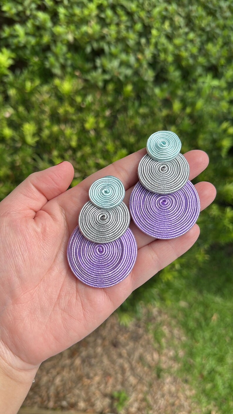 Pode incluir: Um par de brincos pendentes com tr&ecirc;s discos circulares empilhados de tamanhos variados. Cada disco &eacute; constru&iacute;do com fio enrolado firmemente em azul claro, prata e roxo, dispostos num padr&atilde;o vertical do menor para o maior.
