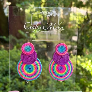 Pode incluir: Um par de brincos composto por tr&ecirc;s elementos circulares de cord&atilde;o soutache empilhados em cores vibrantes de arco-&iacute;ris, incluindo rosa, roxo, azul e amarelo. Os brincos est&atilde;o expostos num suporte de acr&iacute;lico transparente com o texto Crafty Moka.