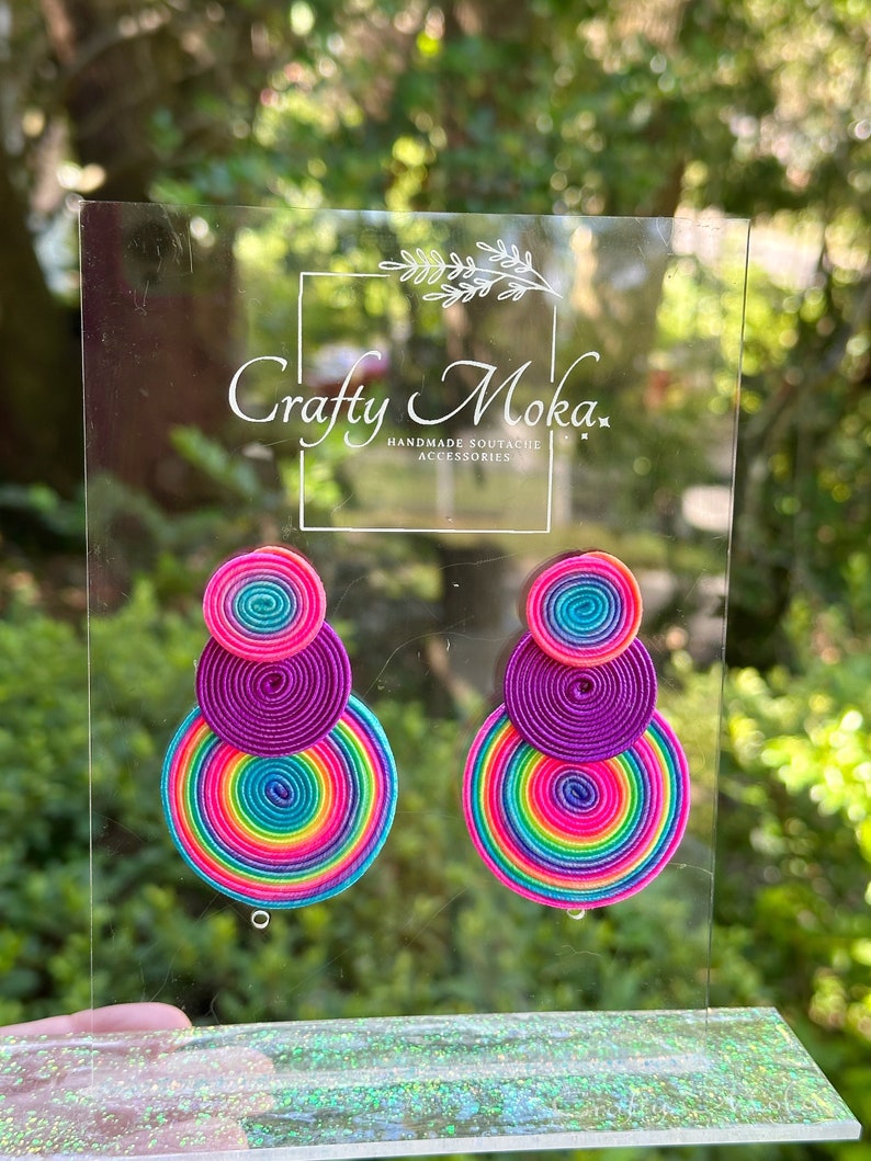 Pode incluir: Um par de brincos composto por tr&ecirc;s elementos circulares de cord&atilde;o soutache empilhados em cores vibrantes de arco-&iacute;ris, incluindo rosa, roxo, azul e amarelo. Os brincos est&atilde;o expostos num suporte de acr&iacute;lico transparente com o texto Crafty Moka.