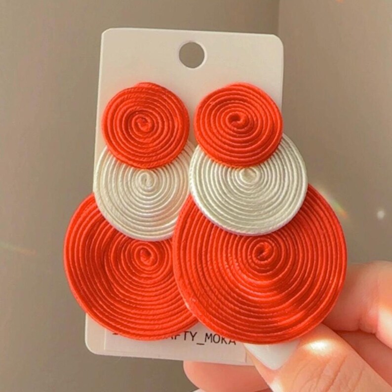 Pode incluir: Um par de brincos pendentes com tr&ecirc;s discos circulares sobrepostos feitos de fio enrolado. Os discos superior e inferior s&atilde;o laranja brilhante, enquanto o disco central &eacute; branco. Fixados num cart&atilde;o branco com o texto CRAFTY_MOKA.
