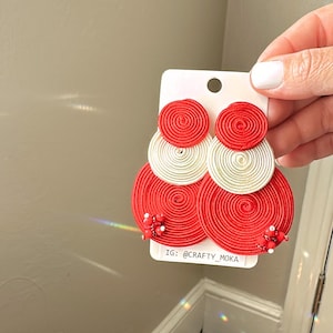 Pode incluir: Um par de brincos circulares em camadas feitos com discos de fio enrolado em vermelho e branco. Os discos vermelhos maiores s&atilde;o decorados com pequenas contas vermelhas e brancas na parte inferior. Est&atilde;o presos a um cart&atilde;o branco com o texto IG: @CRAFTY_MOKA.