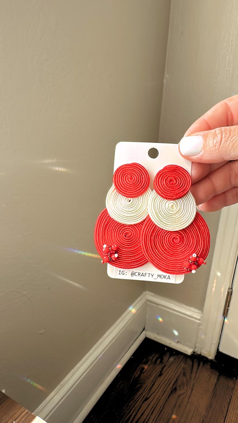 Pode incluir: Um par de brincos circulares em camadas feitos com discos de fio enrolado em vermelho e branco. Os discos vermelhos maiores s&atilde;o decorados com pequenas contas vermelhas e brancas na parte inferior. Est&atilde;o presos a um cart&atilde;o branco com o texto IG: @CRAFTY_MOKA.