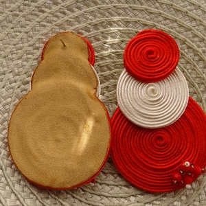 Pode incluir: Dois brincos circulares com bobinas conc&ecirc;ntricas de fio vermelho e branco. Um brinco mostra um verso de couro liso na cor castanho-claro, enquanto o outro exibe a frente com espirais de fio e pequenas contas vermelhas e brancas.