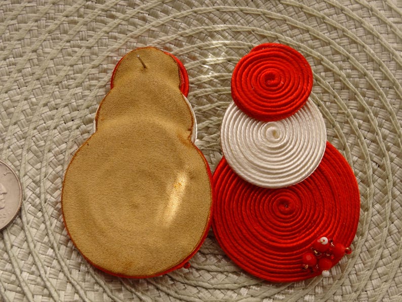 Pode incluir: Dois brincos circulares com bobinas conc&ecirc;ntricas de fio vermelho e branco. Um brinco mostra um verso de couro liso na cor castanho-claro, enquanto o outro exibe a frente com espirais de fio e pequenas contas vermelhas e brancas.