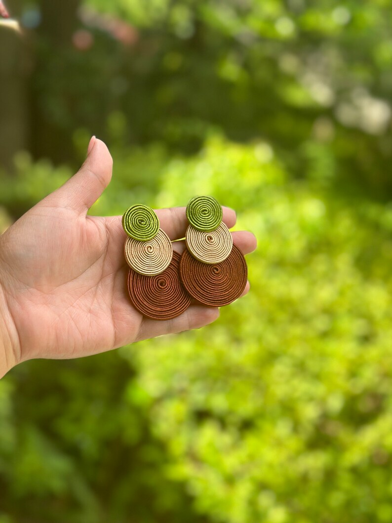 Pode incluir: Um par de brincos pendentes com tr&ecirc;s discos circulares sobrepostos feitos de fio enrolado. O disco superior &eacute; verde-lim&atilde;o, o disco do meio &eacute; bege e o disco inferior &eacute; castanho-escuro. Cada brinco &eacute; composto por estes tr&ecirc;s c&iacute;rculos coloridos.