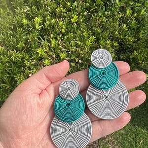 Pode incluir: Um par de brincos com tr&ecirc;s discos circulares sobrepostos de tamanhos variados. Os discos s&atilde;o feitos de fio met&aacute;lico prateado e azul-petr&oacute;leo enrolado, criando um padr&atilde;o espiral texturizado com um design vertical.