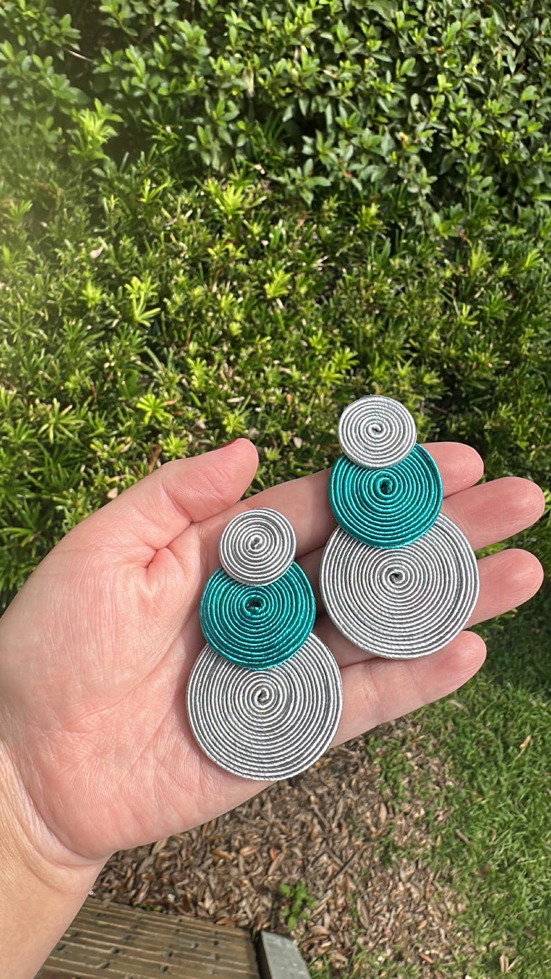 Pode incluir: Um par de brincos com tr&ecirc;s discos circulares sobrepostos de tamanhos variados. Os discos s&atilde;o feitos de fio met&aacute;lico prateado e azul-petr&oacute;leo enrolado, criando um padr&atilde;o espiral texturizado com um design vertical.