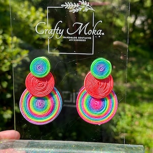 Pode incluir: Um par de brincos com tr&ecirc;s componentes circulares empilhados feitos de cord&atilde;o enrolado em cores vibrantes de arco-&iacute;ris, incluindo verde neon, vermelho, amarelo e roxo. Os brincos est&atilde;o num suporte de acr&iacute;lico transparente com o texto Crafty Moka.