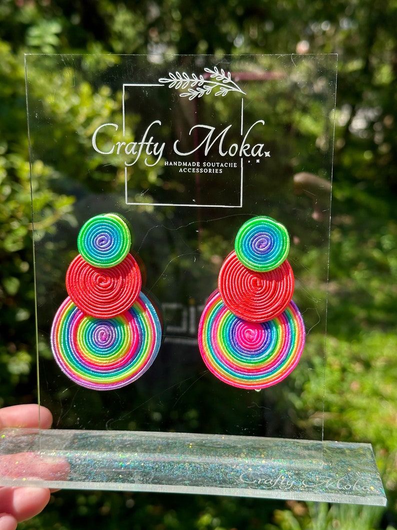 Pode incluir: Um par de brincos com tr&ecirc;s componentes circulares empilhados feitos de cord&atilde;o enrolado em cores vibrantes de arco-&iacute;ris, incluindo verde neon, vermelho, amarelo e roxo. Os brincos est&atilde;o num suporte de acr&iacute;lico transparente com o texto Crafty Moka.