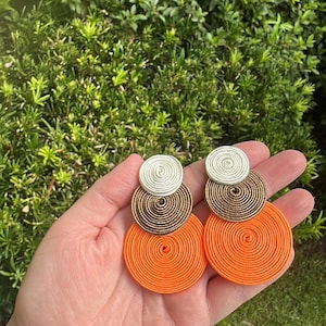 Pode incluir: Um par de brincos pendentes com tr&ecirc;s discos circulares sobrepostos de tamanhos variados. O disco superior &eacute; branco, o disco do meio &eacute; bronze e o disco inferior &eacute; laranja brilhante. Cada disco &eacute; feito de fio ou cord&atilde;o enrolado.