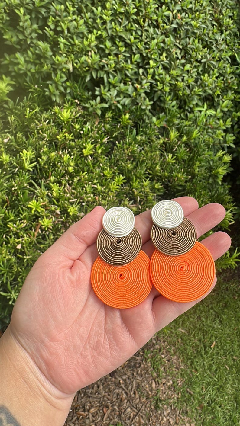 Pode incluir: Um par de brincos pendentes com tr&ecirc;s discos circulares sobrepostos de tamanhos variados. O disco superior &eacute; branco, o disco do meio &eacute; bronze e o disco inferior &eacute; laranja brilhante. Cada disco &eacute; feito de fio ou cord&atilde;o enrolado.