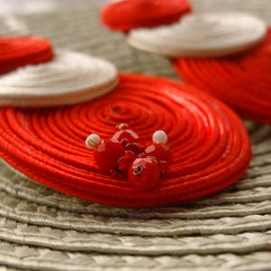 Pode incluir: Um par de brincos circulares com bobinas conc&ecirc;ntricas de fio vermelho e branco. A bobina vermelha maior &eacute; acentuada com um grupo de pequenas contas vermelhas facetadas e contas brancas min&uacute;sculas no centro, criando um visual texturizado.