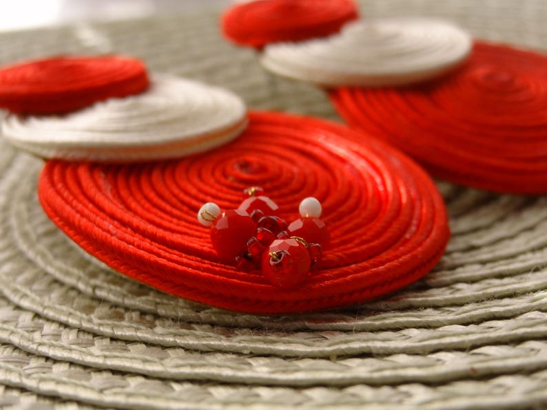 Pode incluir: Um par de brincos circulares com bobinas conc&ecirc;ntricas de fio vermelho e branco. A bobina vermelha maior &eacute; acentuada com um grupo de pequenas contas vermelhas facetadas e contas brancas min&uacute;sculas no centro, criando um visual texturizado.