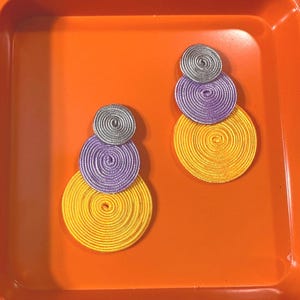 Pode incluir: Um par de brincos com tr&ecirc;s discos circulares empilhados em tamanhos graduados. Cada disco &eacute; constru&iacute;do com fio enrolado nas cores prata, roxo e amarelo. Os brincos est&atilde;o dispostos verticalmente com o disco menor no topo e o maior na parte inferior.