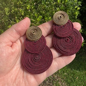 Pode incluir: Um par de brincos com um design empilhado de componentes circulares. Cada brinco consiste num pequeno disco em espiral de cor dourada no topo, seguido por dois discos em espiral maiores de cor bord&ocirc; escuro num padr&atilde;o vertical.