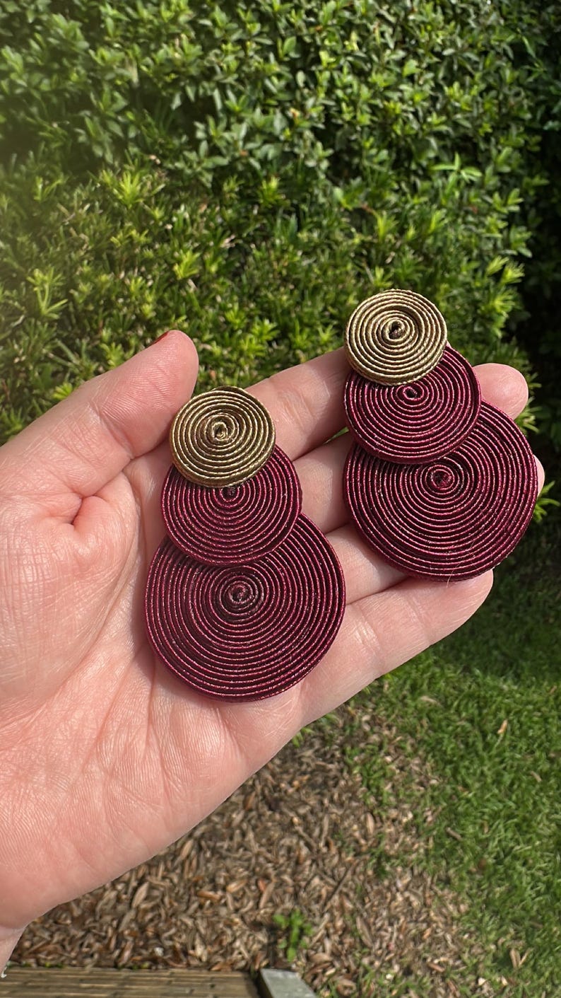 Pode incluir: Um par de brincos com um design empilhado de componentes circulares. Cada brinco consiste num pequeno disco em espiral de cor dourada no topo, seguido por dois discos em espiral maiores de cor bord&ocirc; escuro num padr&atilde;o vertical.