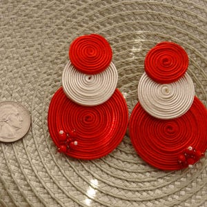 Pode incluir: Um par de brincos pendentes com tr&ecirc;s discos circulares sobrepostos feitos de cord&atilde;o vermelho e branco enrolado. Os discos inferiores maiores s&atilde;o decorados com pequenas contas vermelhas e brancas. Uma moeda de 25 centavos serve de escala.