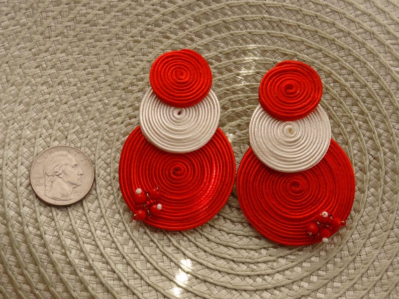 Pode incluir: Um par de brincos pendentes com tr&ecirc;s discos circulares sobrepostos feitos de cord&atilde;o vermelho e branco enrolado. Os discos inferiores maiores s&atilde;o decorados com pequenas contas vermelhas e brancas. Uma moeda de 25 centavos serve de escala.