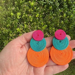 Pode incluir: Um par de brincos pendentes com tr&ecirc;s discos circulares sobrepostos em tamanhos graduados. Cada disco &eacute; constru&iacute;do com fio enrolado firmemente em tons vibrantes de rosa choque, turquesa brilhante e laranja n&eacute;on, criando um design geom&eacute;trico em camadas.