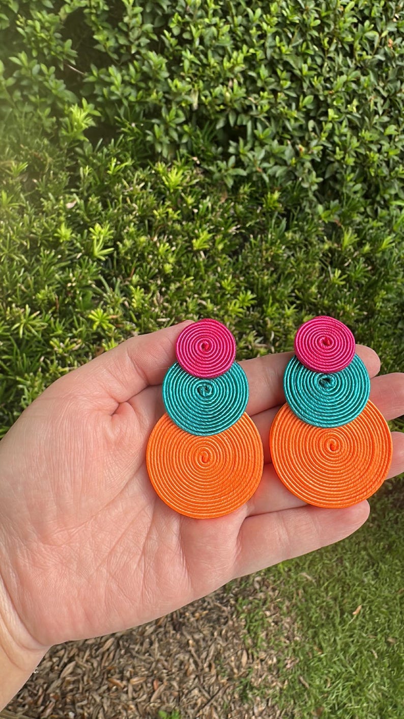 Pode incluir: Um par de brincos pendentes com tr&ecirc;s discos circulares sobrepostos em tamanhos graduados. Cada disco &eacute; constru&iacute;do com fio enrolado firmemente em tons vibrantes de rosa choque, turquesa brilhante e laranja n&eacute;on, criando um design geom&eacute;trico em camadas.