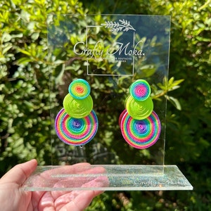 Pode incluir: Um par de brincos com tr&ecirc;s discos circulares empilhados em rosa n&eacute;on, amarelo e um padr&atilde;o de espiral arco-&iacute;ris multicolorido. Os brincos est&atilde;o expostos num suporte de acr&iacute;lico transparente com o texto Crafty Moka Handmade Soutache Accessories.