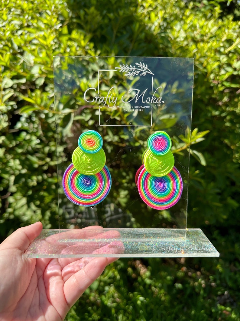 Pode incluir: Um par de brincos com tr&ecirc;s discos circulares empilhados em rosa n&eacute;on, amarelo e um padr&atilde;o de espiral arco-&iacute;ris multicolorido. Os brincos est&atilde;o expostos num suporte de acr&iacute;lico transparente com o texto Crafty Moka Handmade Soutache Accessories.