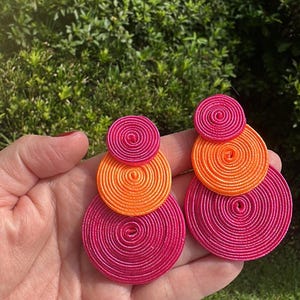Pode incluir: Um par de brincos com discos circulares empilhados feitos de fio enrolado. O brinco esquerdo consiste num pequeno disco rosa choque, um disco laranja m&eacute;dio e um grande disco rosa choque. O brinco direito apresenta um pequeno disco rosa choque, um disco laranja m&eacute;dio e um grande disco rosa choque.