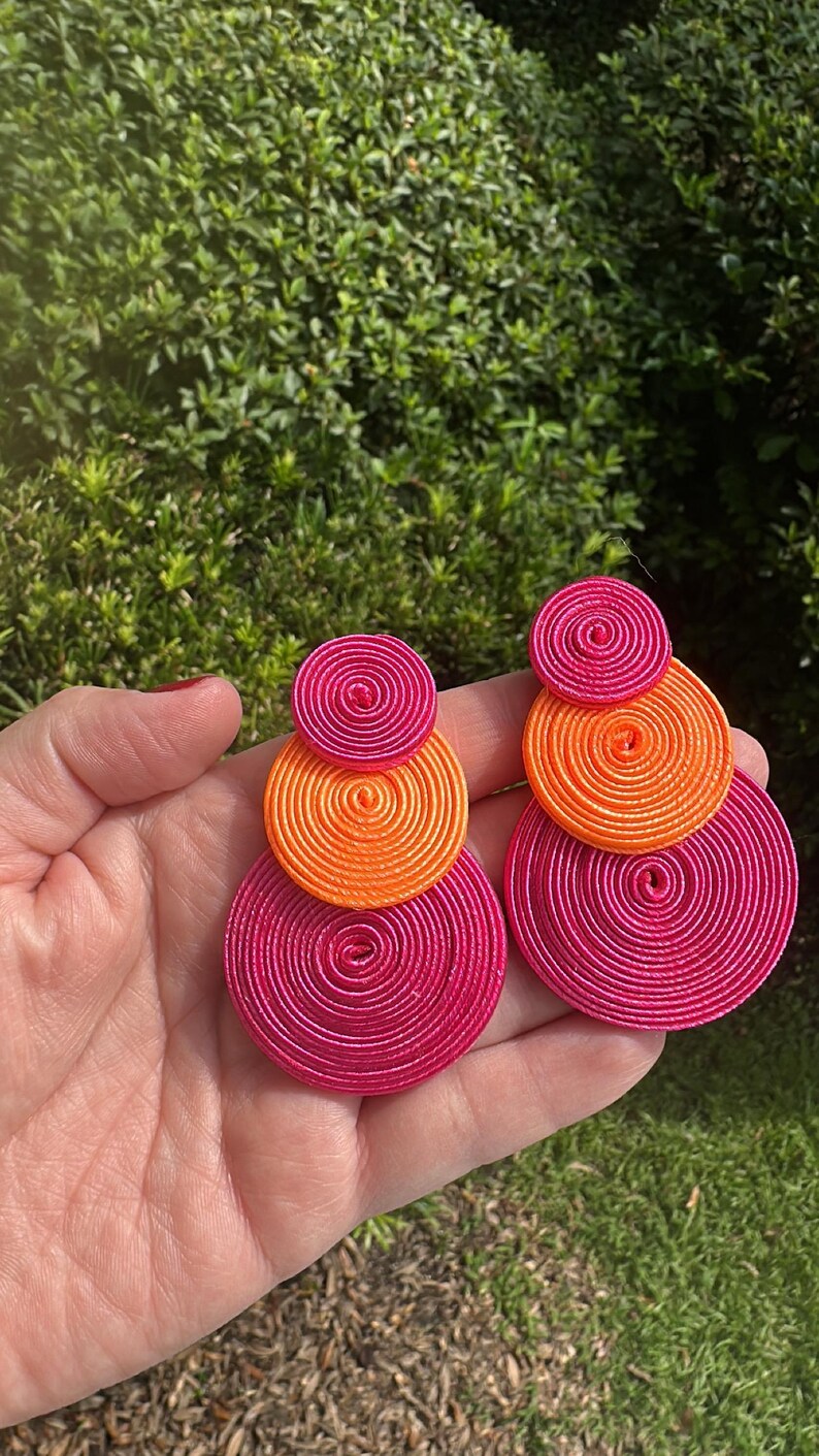Pode incluir: Um par de brincos com discos circulares empilhados feitos de fio enrolado. O brinco esquerdo consiste num pequeno disco rosa choque, um disco laranja m&eacute;dio e um grande disco rosa choque. O brinco direito apresenta um pequeno disco rosa choque, um disco laranja m&eacute;dio e um grande disco rosa choque.