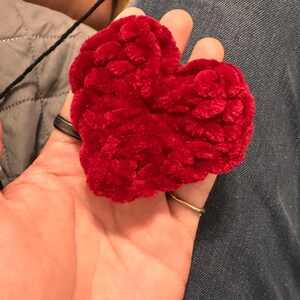 May include: A deep red, heart-shaped item made from a soft, textured material. The item is held in a hand, with a black string attached. The item could be a decorative piece or a small cushion.