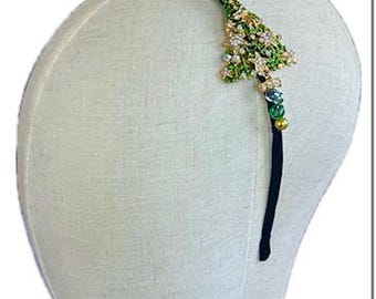 May include: A thin black headband featuring a decorative Christmas tree ornament made of green and gold metal, accented with small clear crystals and various green and gold beads arranged along the band to create a festive holiday accessory.