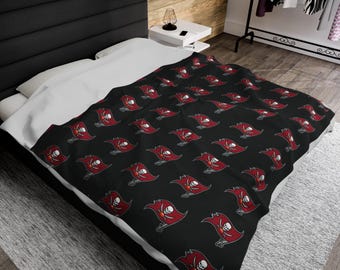 May include: A black fleece blanket featuring a repeating pattern of the Tampa Bay Buccaneers logo, which consists of a red pirate flag with a white skull and crossbones design, draped over a bed with white bedding and a black headboard.