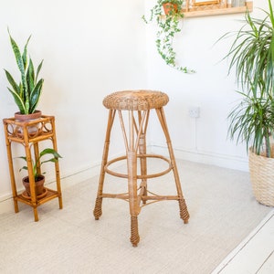 May include: A wicker stool with a round seat and a woven design. The stool is on a white rug in a room with white walls and plants.