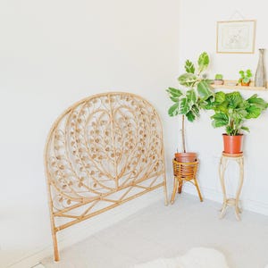 Könnte beinhalten: Ein natürliches Kopfteil aus Rattan mit gewölbter Oberseite und dekorativem Blumendesign. Das Kopfteil steht vor einer weißen Wand, mit Pflanzen in Töpfen und einem kleinen Regal mit Dekoration im Hintergrund.