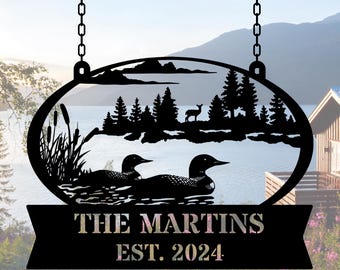 May include: Black metal sign featuring a lake scene with two loons, trees, and a deer silhouette. The sign is oval-shaped with a banner below that reads "THE MARTINS EST. 2024". It hangs from chains, suitable for outdoor display.
