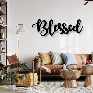 May include: Black metal wall art spelling out the word "Blessed" in a cursive font. The sign is mounted on a white wall above a brown leather sofa with decorative pillows. The room is decorated with a floor lamp, side tables, and a woven rug.