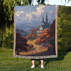 May include: A woven throw blanket featuring a landscape painting of a castle on a hill with autumn trees. The artwork depicts a cloudy sky and a path leading to the castle. The blanket has a fringed edge.
