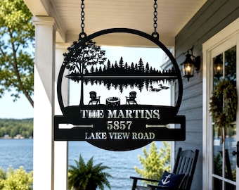 May include: A black metal hanging sign featuring a circular frame with a silhouette of a lake, trees, two chairs, a fire pit, and ducks. The text reads THE MARTINS 5857 LAKE VIEW ROAD, accented by two horizontal oar icons on the lower rectangular panel.