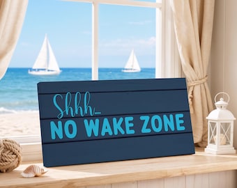 May include: A navy blue wooden sign with the text "Shhh... NO WAKE ZONE" in turquoise. The sign is placed on a wooden windowsill with a beach scene visible through a window in the background.