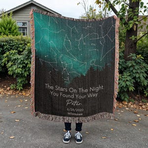 May include: A woven throw blanket featuring a celestial design with constellations and a gradient of teal and black. The blanket has a fringed edge and includes the text "The Stars On The Night You Found Your Way" with a name, date, and city.