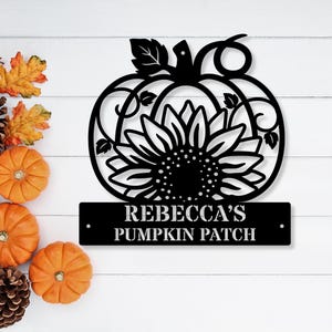 May include: Black metal pumpkin sign with a sunflower design and the text "REBECCA'S PUMPKIN PATCH". The sign is decorated with a pumpkin and sunflower design, perfect for fall decor.