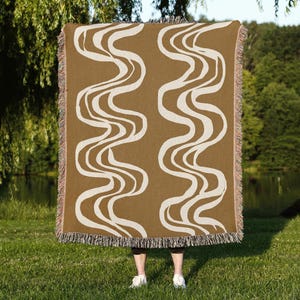 May include: A brown woven throw blanket with a cream-colored wavy pattern. The blanket has a fringed edge and is held up in front of a person standing on grass. The background includes a lake and trees.