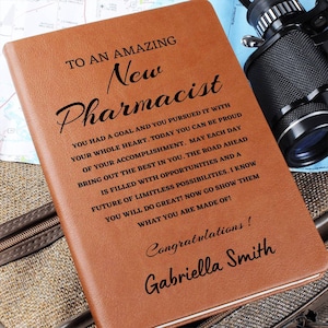 May include: A brown leather journal with the text "To an amazing new Pharmacist" and a congratulatory message for Gabriella Smith. The journal is a gift for a new pharmacist.